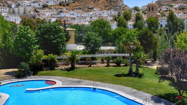 A swimming pool with a clear blue water, surrounded by a paved area with a few trees and a view of a hillside town in the background.