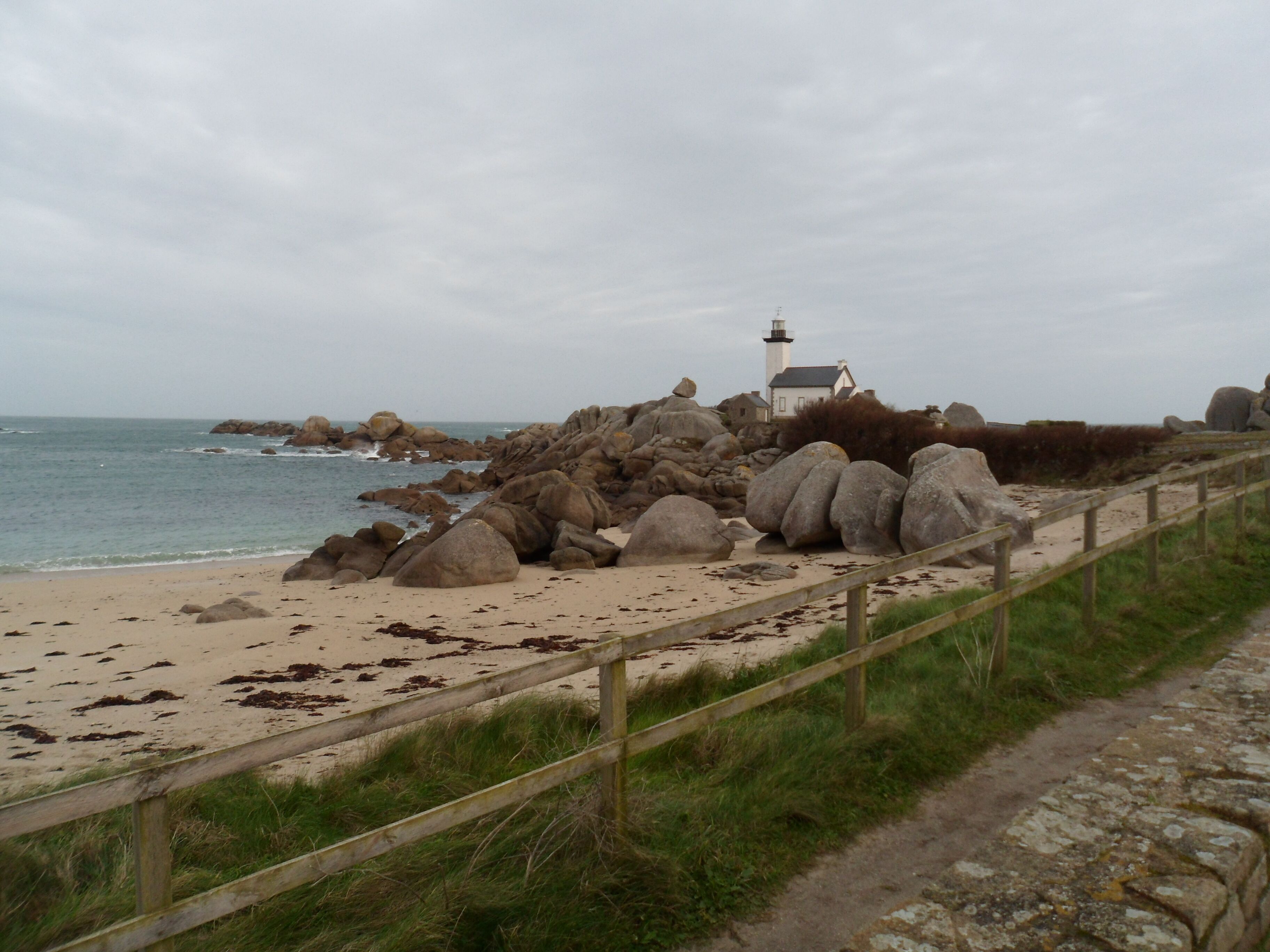 Un sendero costero con una valla de madera, rocas grandes y un faro al fondo.