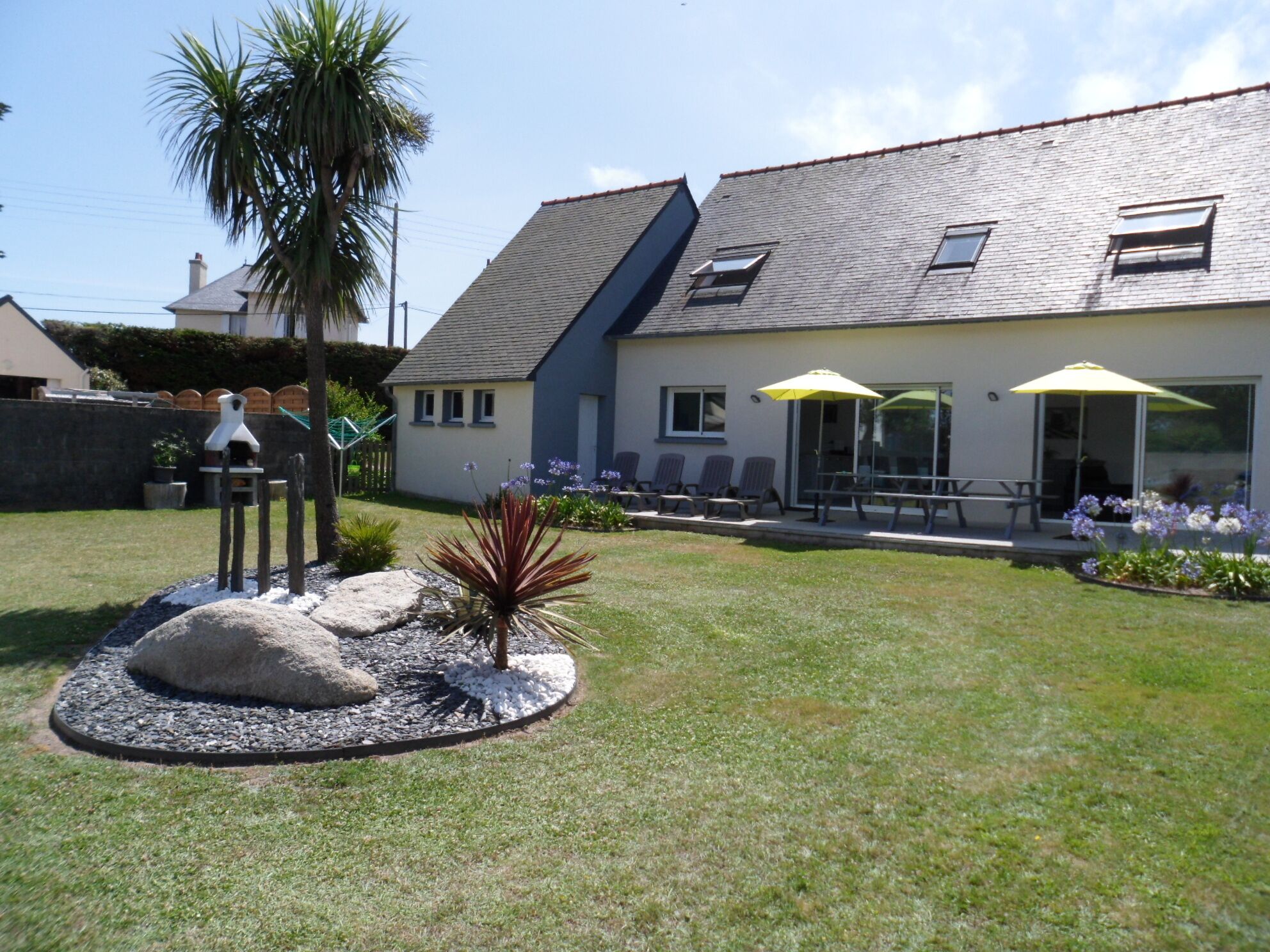 A house with a sloped roof, a patio with chairs and umbrellas, and a garden with a palm tree and plants.
