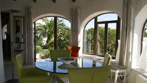 Una habitación con una mesa y sillas de vidrio, ventanas amplias con vistas a palmeras y una pequeña mesa blanca.