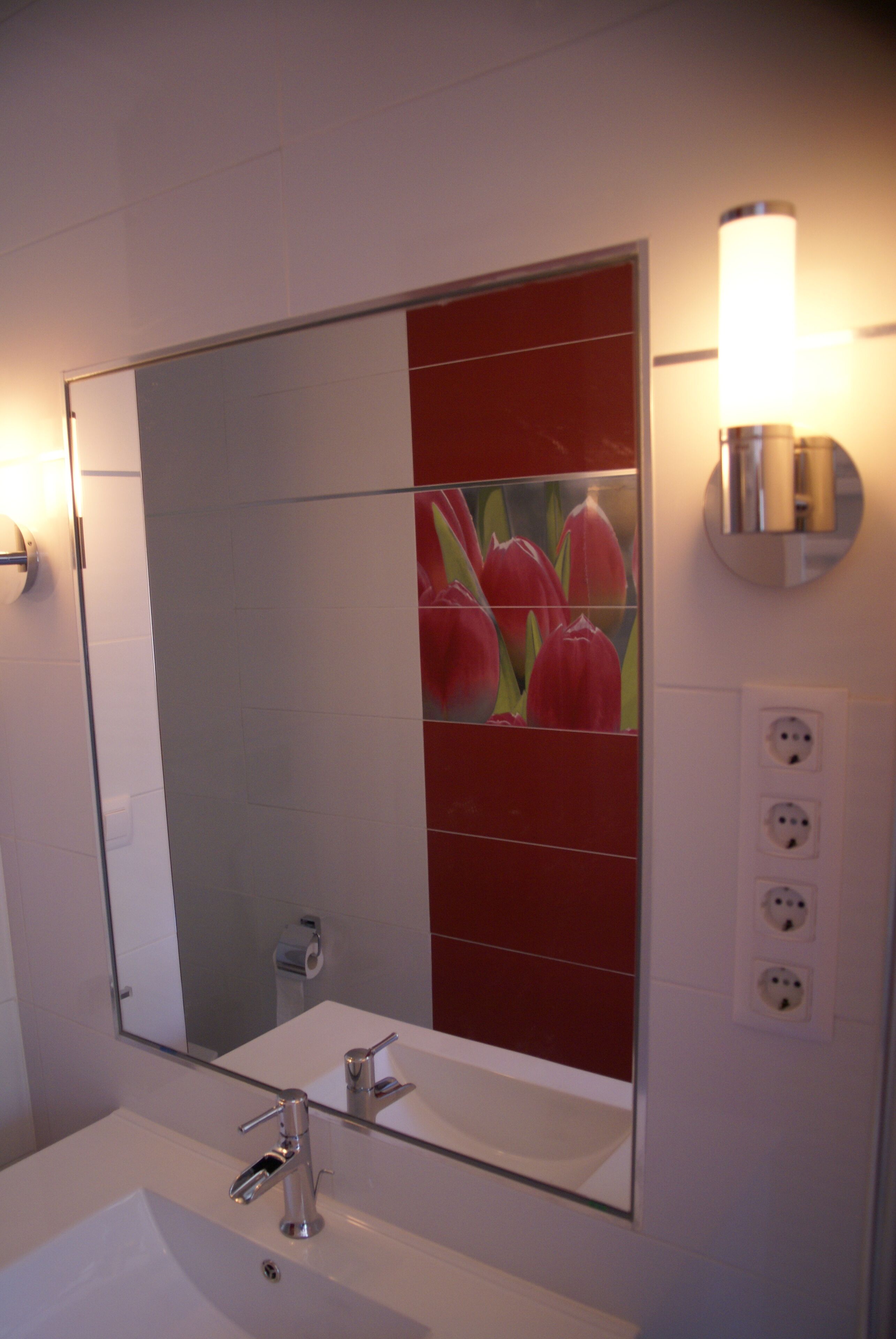 Une salle de bain avec un lavabo blanc, un miroir et une décoration murale à motifs floraux rouges.