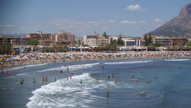 Una playa concurrida, gente nadando y tomando el sol, y edificios al fondo.