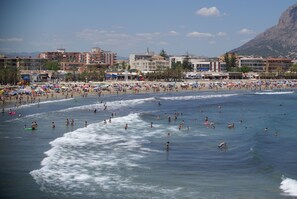 Una playa concurrida, gente nadando y tomando el sol, y edificios al fondo.