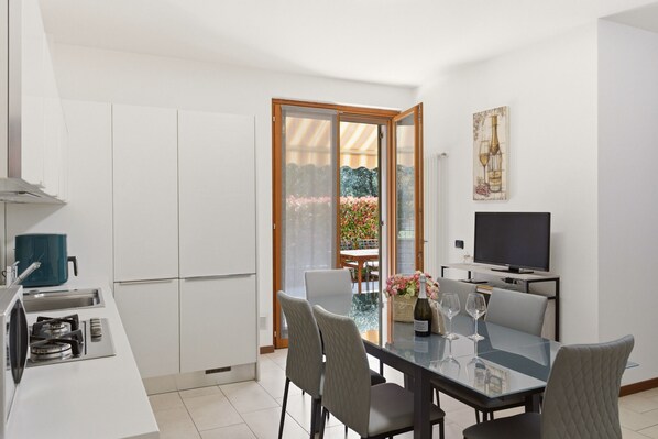 A modern kitchen with a stove, microwave, and a blue container. There is a dining area with a glass table and chairs, a TV, and a painting on the wall.