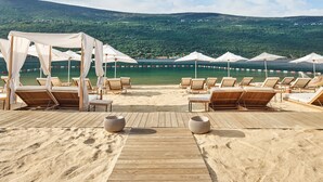 Een terras aan het strand met ligbedden en parasols, met uitzicht op een rustige zee en groene heuvels.