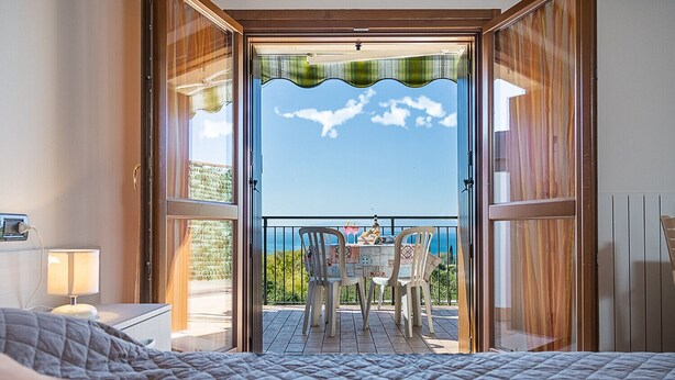 A bedroom with an open door leading to a balcony with a table and chairs, overlooking a scenic view.