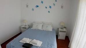 Un letto ben rifatto con una coperta blu e cuscini bianchi, due comodini con lampade, e un muro decorato con sagome di uccelli e lanterne.