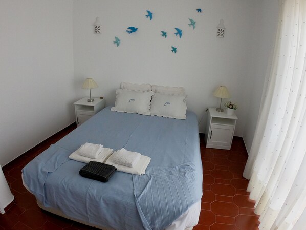 A neatly made bed with a blue bedspread and white pillows, two bedside tables with lamps, and a wall decorated with paper cutouts of birds and lanterns.