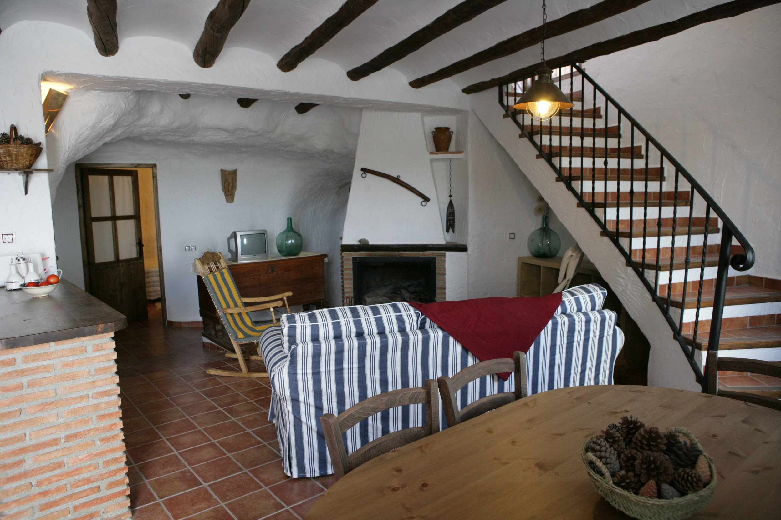 Un interior rústico con vigas de madera a la vista, una chimenea, un área de comedor con tapicería a rayas y una escalera con barandilla de hierro forjado.