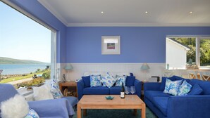 A room with a large window overlooking a scenic view, a blue sofa with patterned cushions, a wooden coffee table, and a small side table with a lamp.