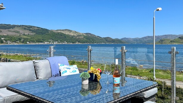 A glass table with a bottle, grapes, a small plant, and a cushion by a lakeside.
