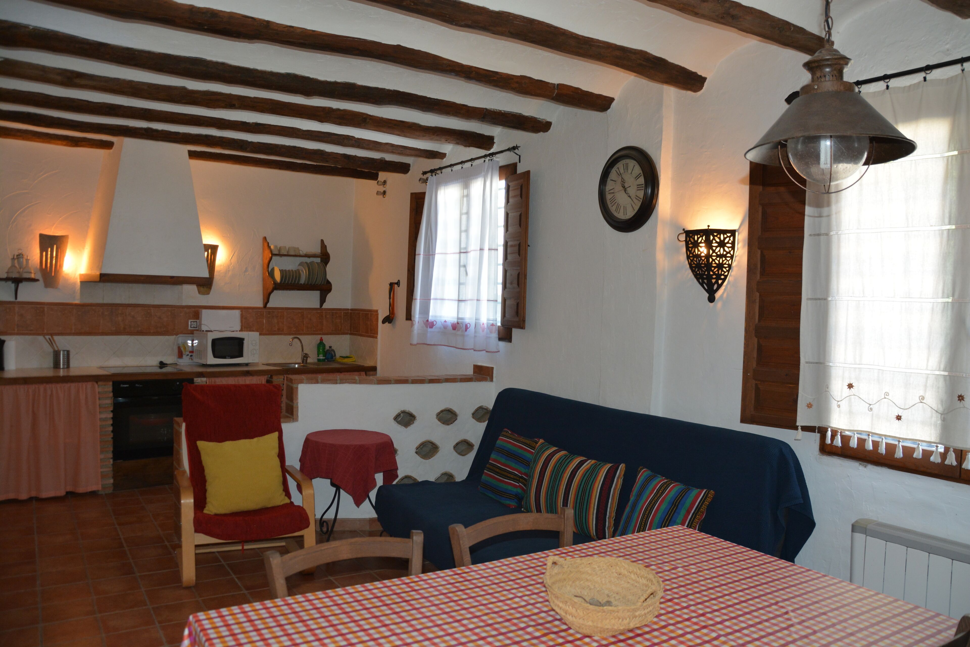 Ein gemütliches Wohnzimmer mit einem karierten Tisch, einem blauen Sofa und einem roten Sessel. Im Hintergrund ist eine Küchenzeile mit einer Mikrowelle und einem Spülbecken zu sehen.