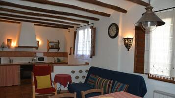 Ein gemütliches Wohnzimmer mit einem karierten Tisch, einem blauen Sofa und einem roten Sessel. Im Hintergrund ist eine Küchenzeile mit einer Mikrowelle und einem Spülbecken zu sehen.