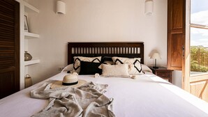 A neatly made bed with a wooden headboard, a hat, and a blanket on the bed. There are two bedside tables with lamps, a window with wooden shutters, and a shelf with decorative items. - CAN NOVES - Detached, central and peaceful villa (Sant Francesc de Formentera)