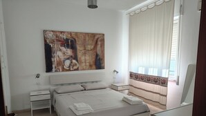 A bedroom with a bed, nightstands, a large abstract painting, a window with curtains, and a mirror. - House with terrace, five minutes from Sottotorre beach (Calasetta)