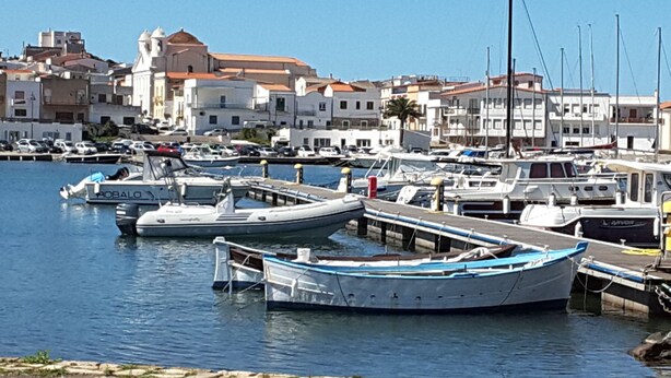 A marina with boats docked, a clear sky, and buildings in the background. - House with terrace, five minutes from Sottotorre beach (Calasetta)