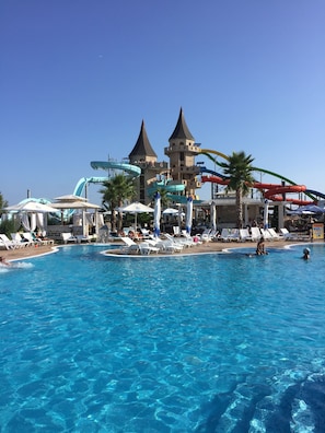A swimming pool with water slides and umbrellas, surrounded by lounge chairs and palm trees.