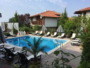 Zona de piscina con sillones de descanso y sombrillas, rodeada de vegetación y un edificio con techo de tejas.
