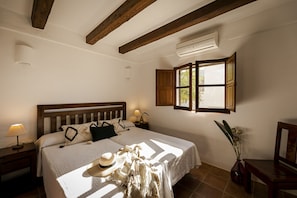 Une chambre avec un plafond en bois, un lit recouvert d’une courtepointe blanche, une table de chevet avec une lampe, une fenêtre avec des volets en bois et un vase rempli de fleurs.