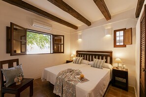 A bedroom with a large bed, wooden headboard, bedside tables with lamps, a nightstand, a chair with a cushion, and a window with a view of trees. - CAN NOVES - Villa 5 suites 9 & 31 (Sant Francesc de Formentera)