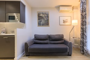 Ein modernes Wohnzimmer mit grauem Sofa, einer Mikrowelle, einem kleinen Tisch mit Lampe und einem Gemälde an der Wand.