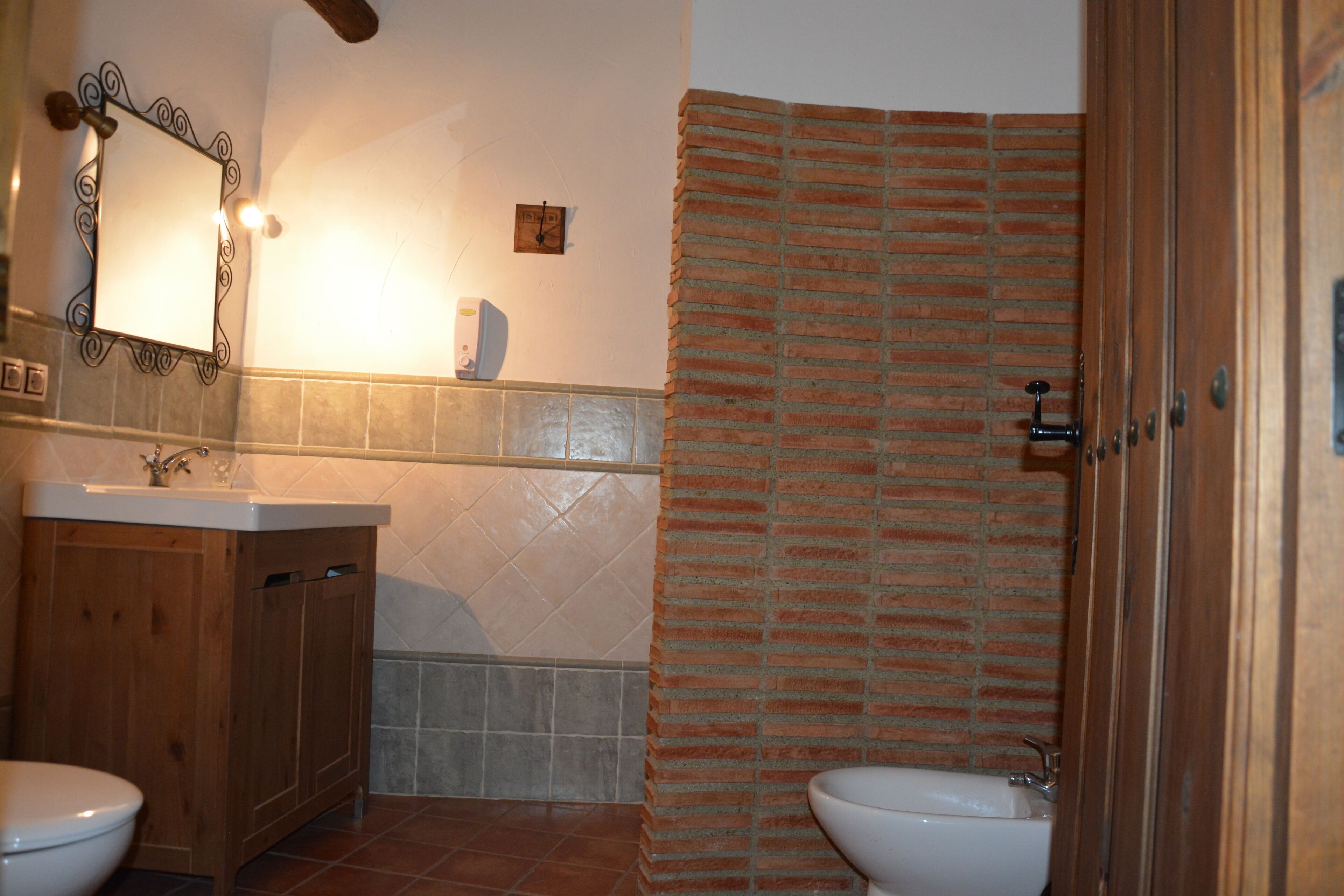 Un baño con un mueble de tocador de madera, un lavabo blanco, un espejo, un inodoro blanco y una ducha con pared de ladrillo.