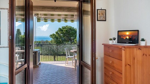 Une porte ouverte donnant sur un balcon avec vue sur la mer, un ordinateur portable posé sur un meuble en bois et un tableau encadré accroché au mur.