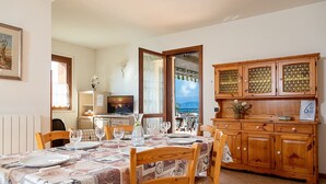 Un coin repas avec une table dressée pour quatre personnes, des meubles en bois et une vue sur la mer par une grande fenêtre.