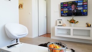 Una sala moderna con un televisor de pantalla plana mostrando un evento deportivo, un sofá blanco, un control remoto, un cuadro enmarcado y un frutero sobre una mesa consola blanca.