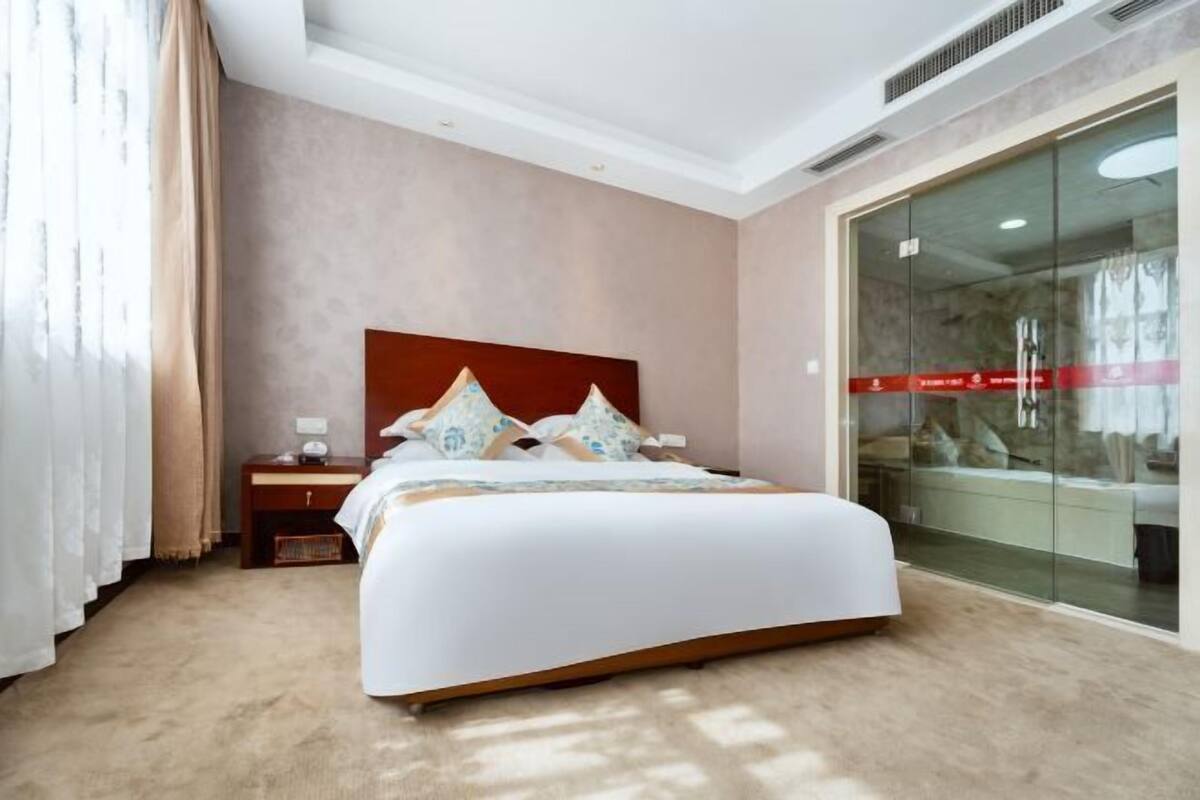 A hotel room with a large bed, a nightstand, a glass partition, and a view of the city.