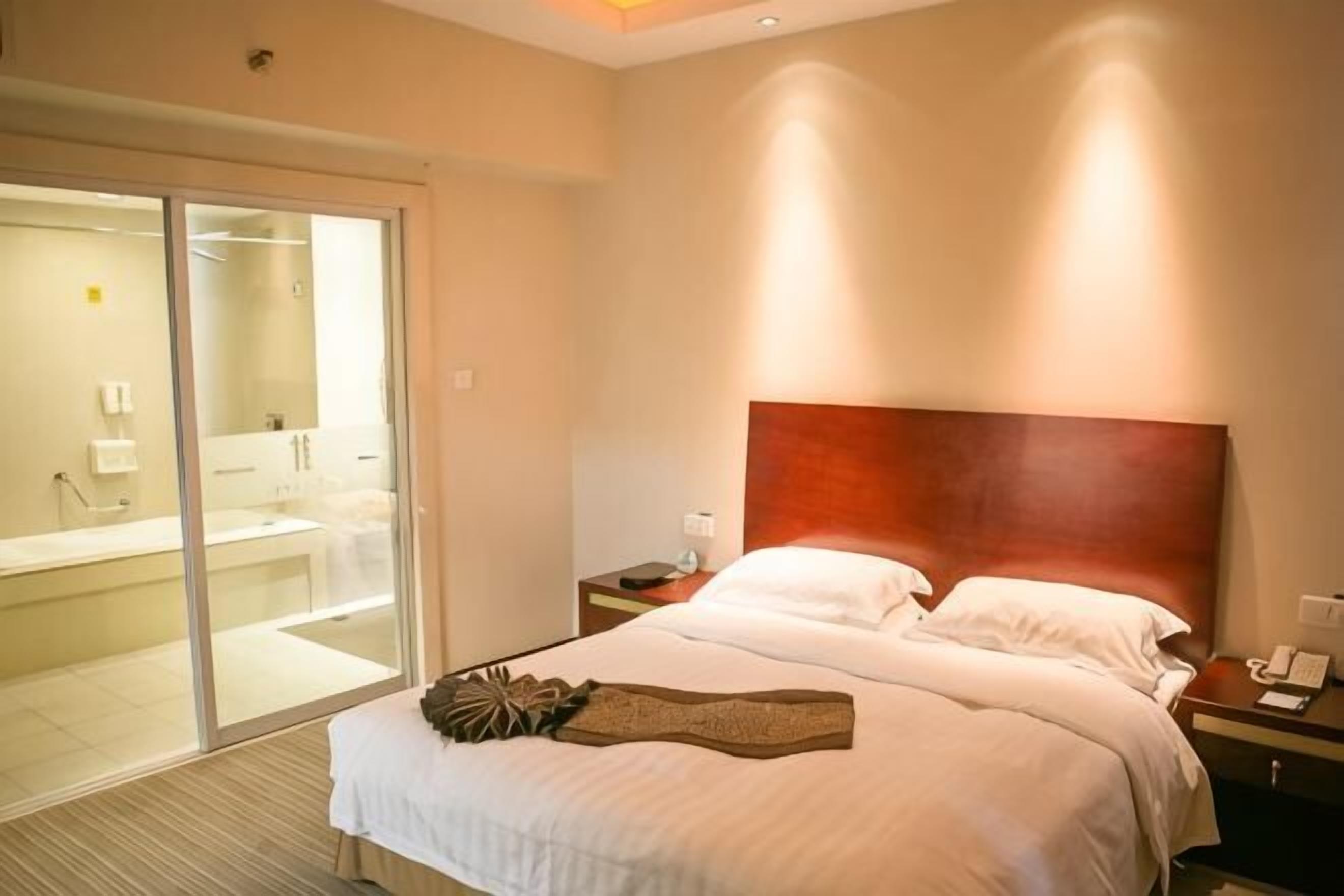 Quarto de hotel com uma cama grande, cabeceira de madeira, mesa de cabeceira e porta de vidro deslizante para um banheiro.