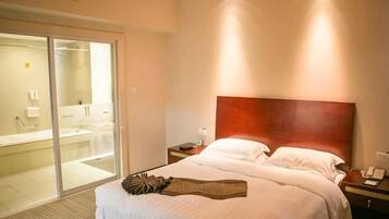 Quarto de hotel com uma cama grande, cabeceira de madeira, mesa de cabeceira e porta de vidro deslizante para um banheiro.