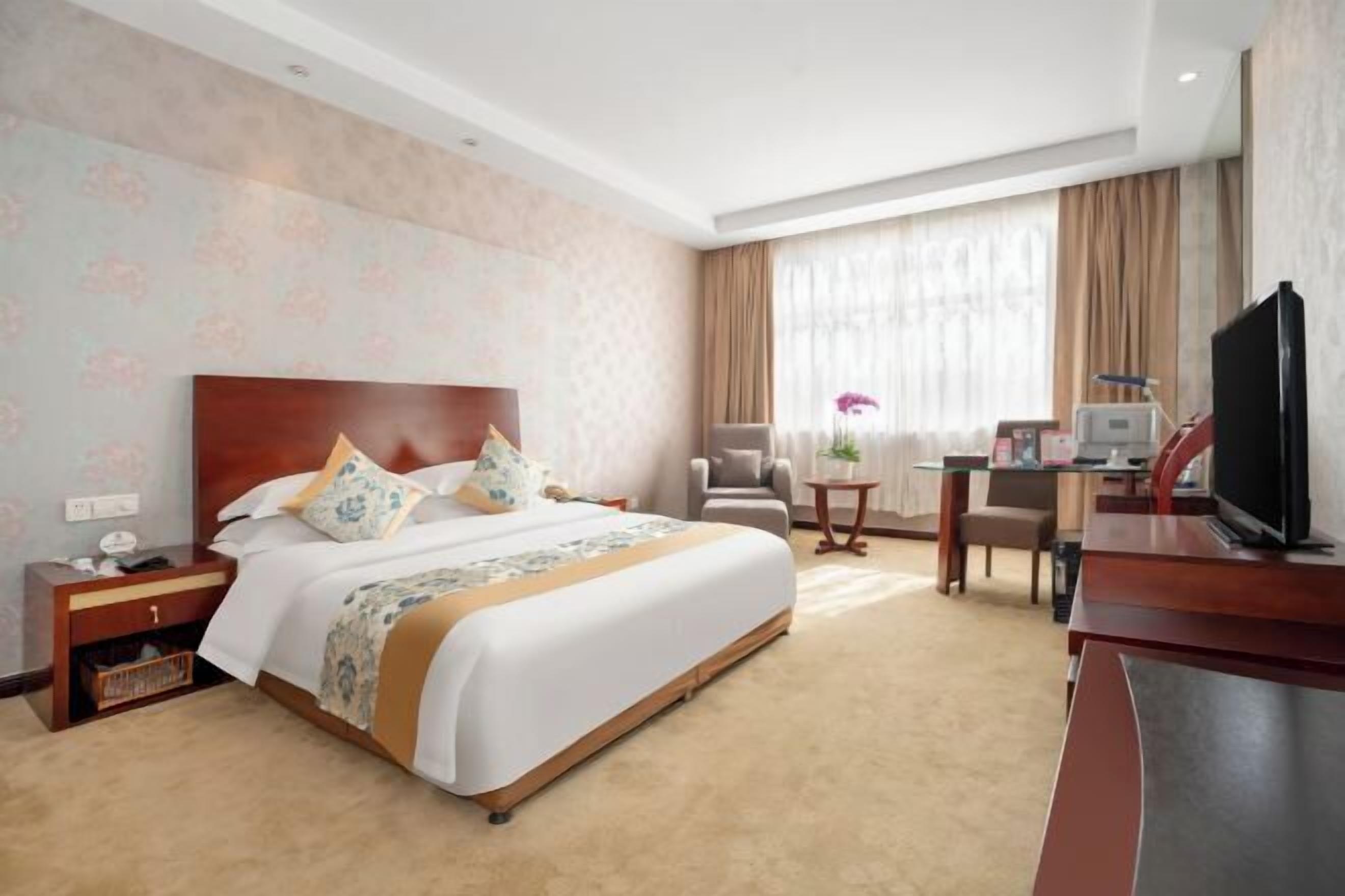 A hotel room with a large bed, a desk with a television, a chair, and a small table with a vase of flowers.