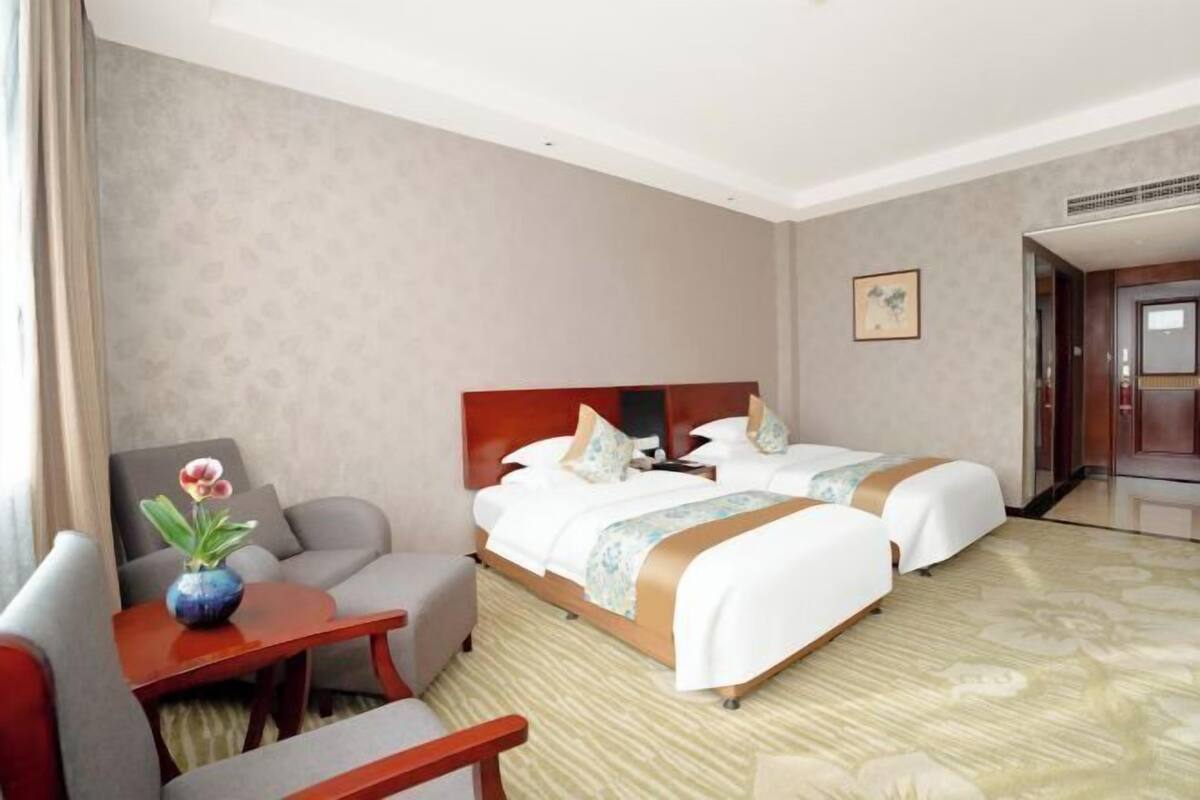 A hotel room with two beds, a sofa, a small table with a vase, and a painting on the wall.
