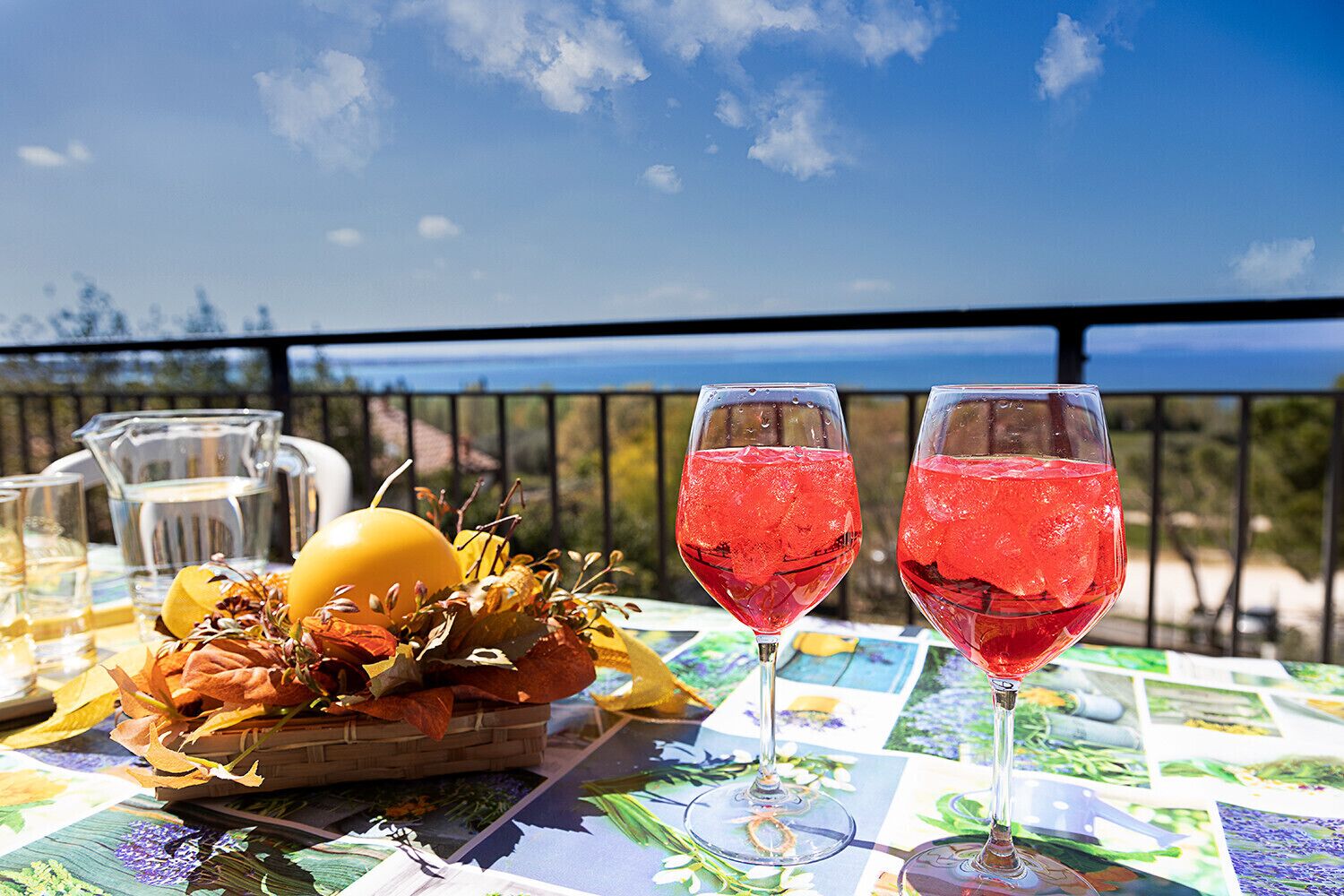 Una mesa con dos vasos de bebida roja, una jarra y una cesta de frutas y hojas en un balcón con vistas al mar.
