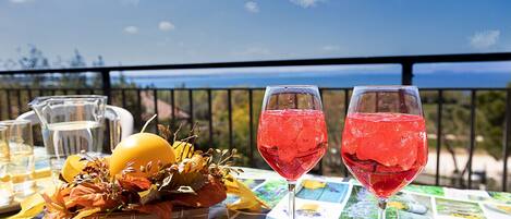 A table with two glasses of red beverage, a pitcher, and a basket of fruits and leaves on a balcony with a view of the sea.