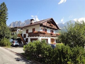 Un chalet à deux étages, doté d’un toit en pente, de balcons en bois ornés de jardinières et surplombant un paysage montagneux.