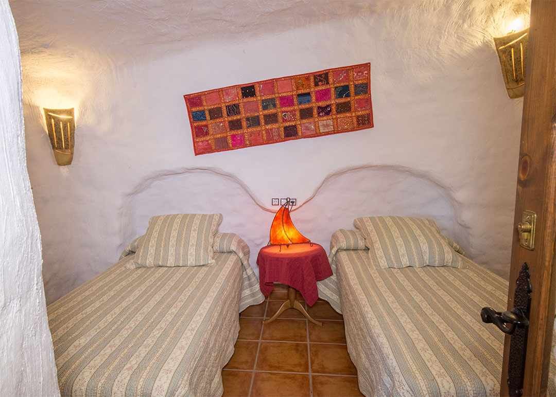 A small, cozy room with two single beds, a red table lamp, and a colorful quilt hanging on the wall.