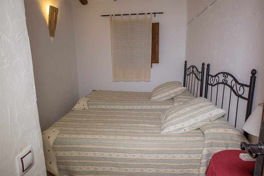 A small, single-bed room with a metal headboard, a bedside table with a lamp, and a window with a curtain.