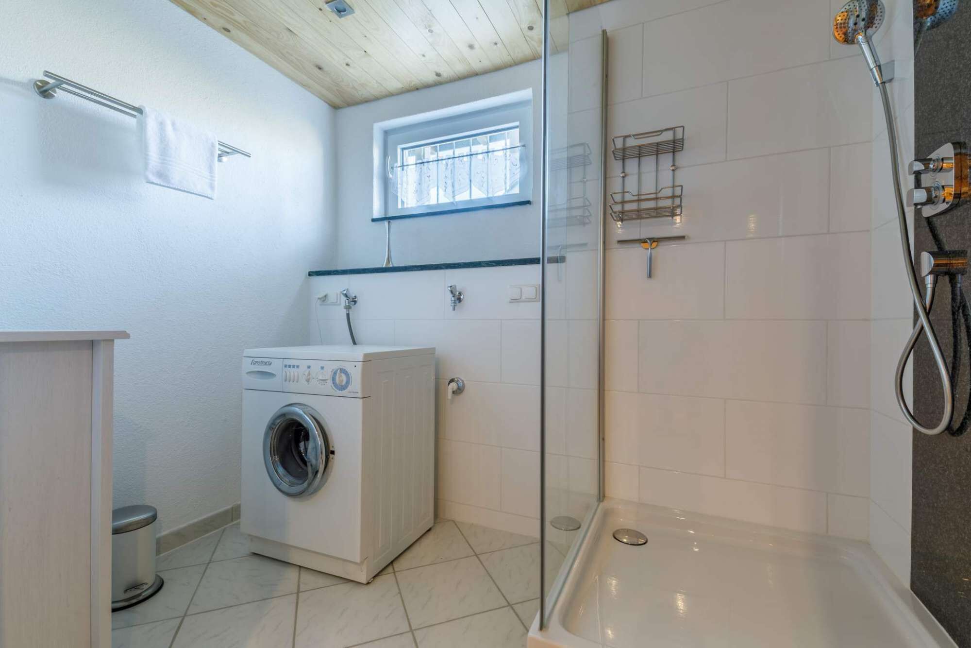 Une salle de bain compacte équipée d’un lave-linge, d’une douche à parois de verre et d’une fenêtre laissant entrer la lumière naturelle.