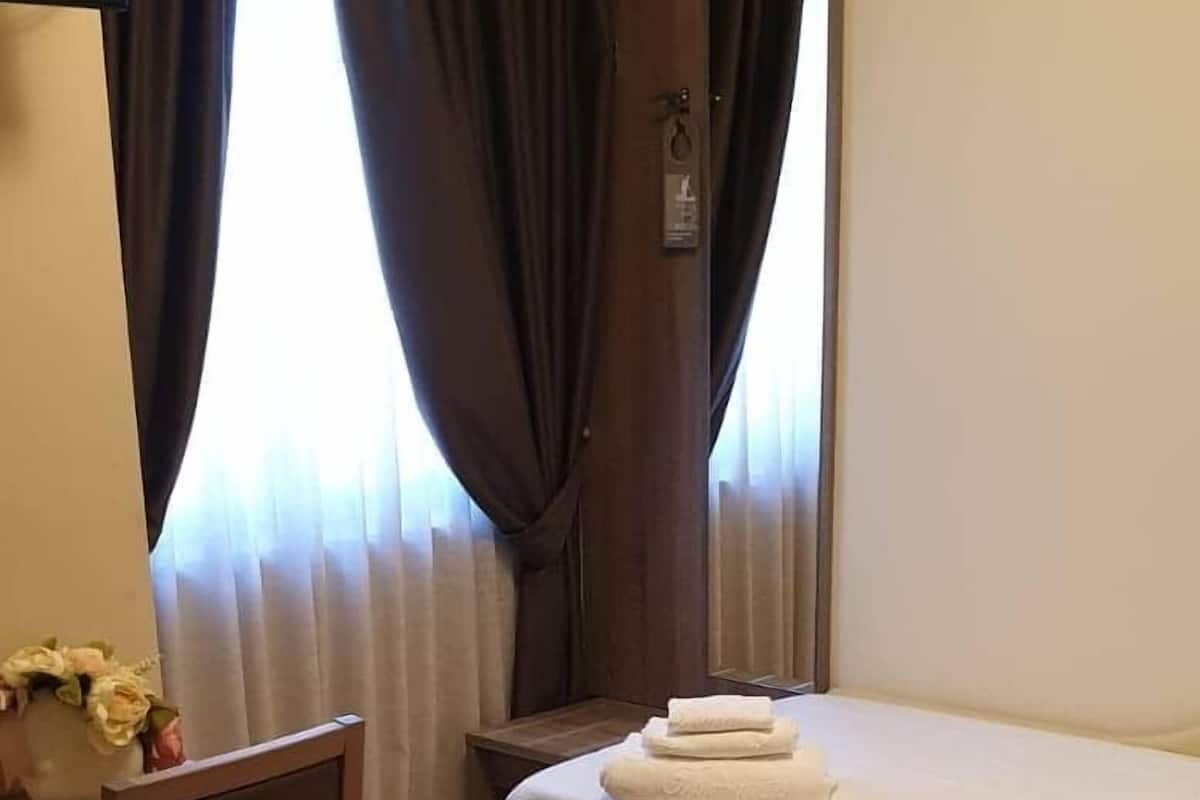 Quarto de hotel com cama, escrivaninha, cadeira e janela com cortinas.