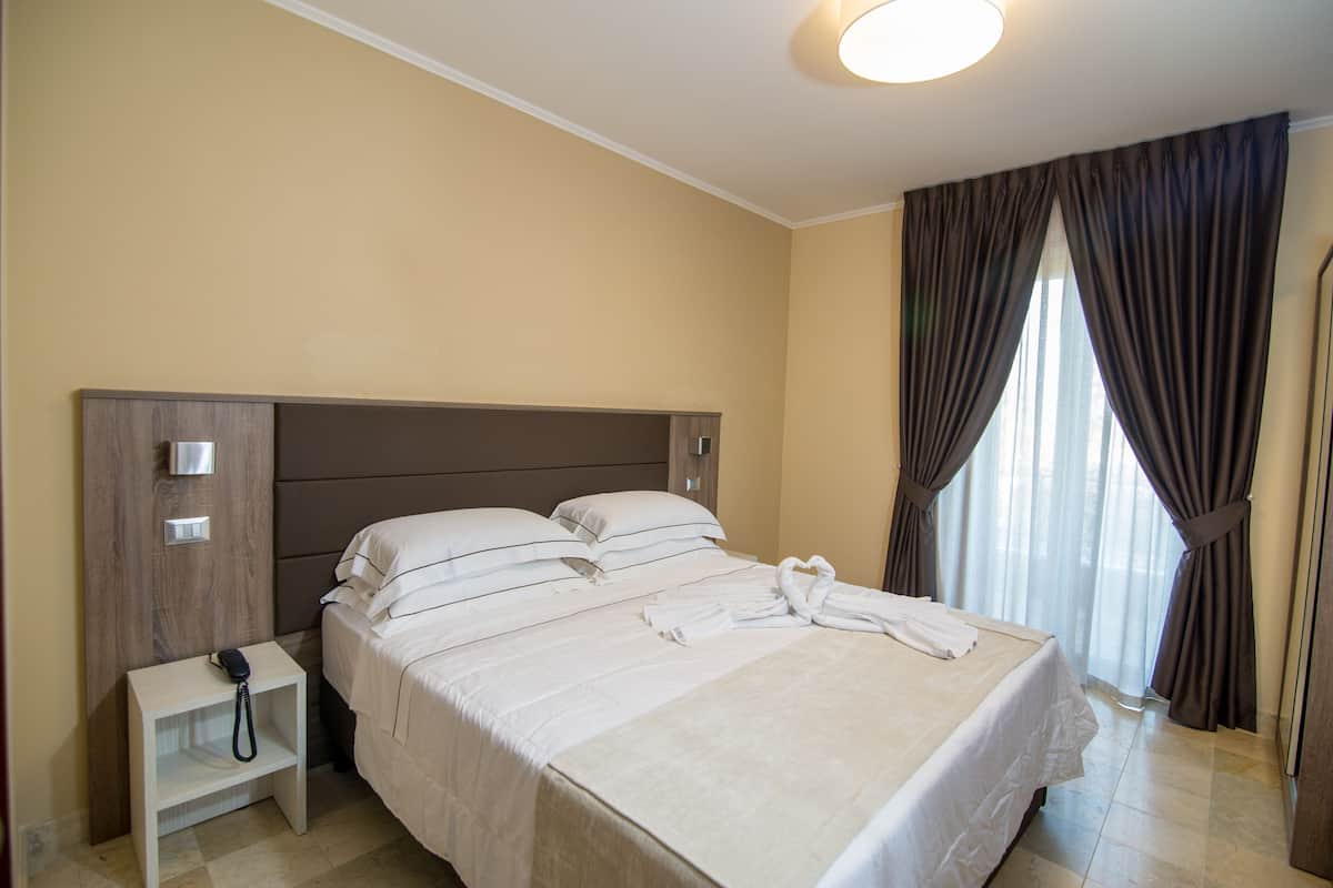 Uma cama bem arrumada com lençóis brancos, uma mesinha de cabeceira com telefone e uma janela com cortinas.