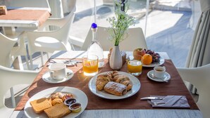 Un desayuno con una mesa preparada para dos personas, que incluye platos de bollería, tostadas y fruta, junto con tazas de café y zumo de naranja.