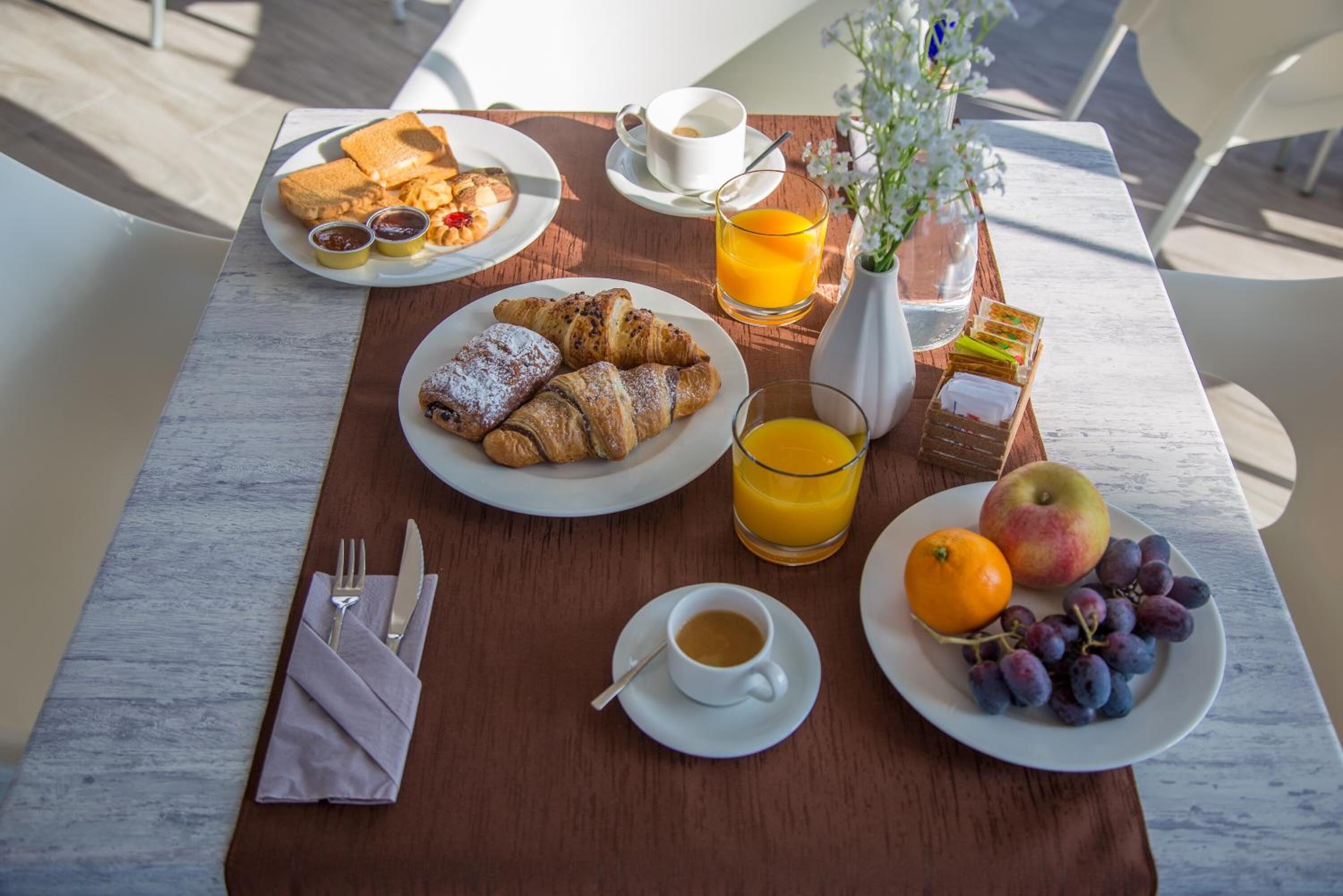 Une table en bois dressée pour le petit-déjeuner, avec des croissants, des fruits, du jus et du café.
