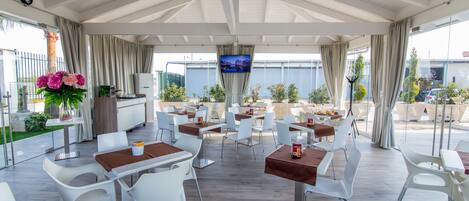 An outdoor dining area with white chairs and tables, a TV screen, and a view of the sea.