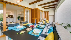 A dining area with a table set for four, a kitchen in the background, and wooden beams on the ceiling.