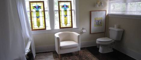 Un baño con dos ventanas de vitral, una silla blanca, un inodoro y un tapete estampado.