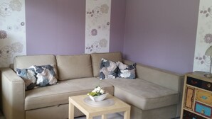 Ein Wohnzimmer mit einem beigefarbenen Sofa, floralen Kissen, einem kleinen Holztisch mit einer Blumenschale und einem Nachttisch im Vintage-Stil mit Schubladen.