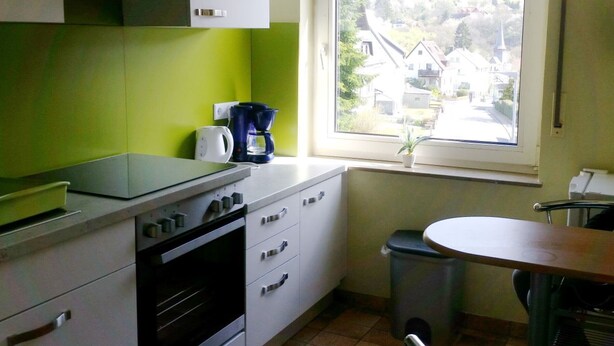 Eine kompakte Küche mit weißen Schränken, einem eingebauten Backofen, einer Kaffeemaschine und einem Fenster mit Blick auf Häuser und Bäume.