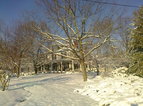 A snow-covered landscape with a house featuring a large porch and a tree in the foreground.
